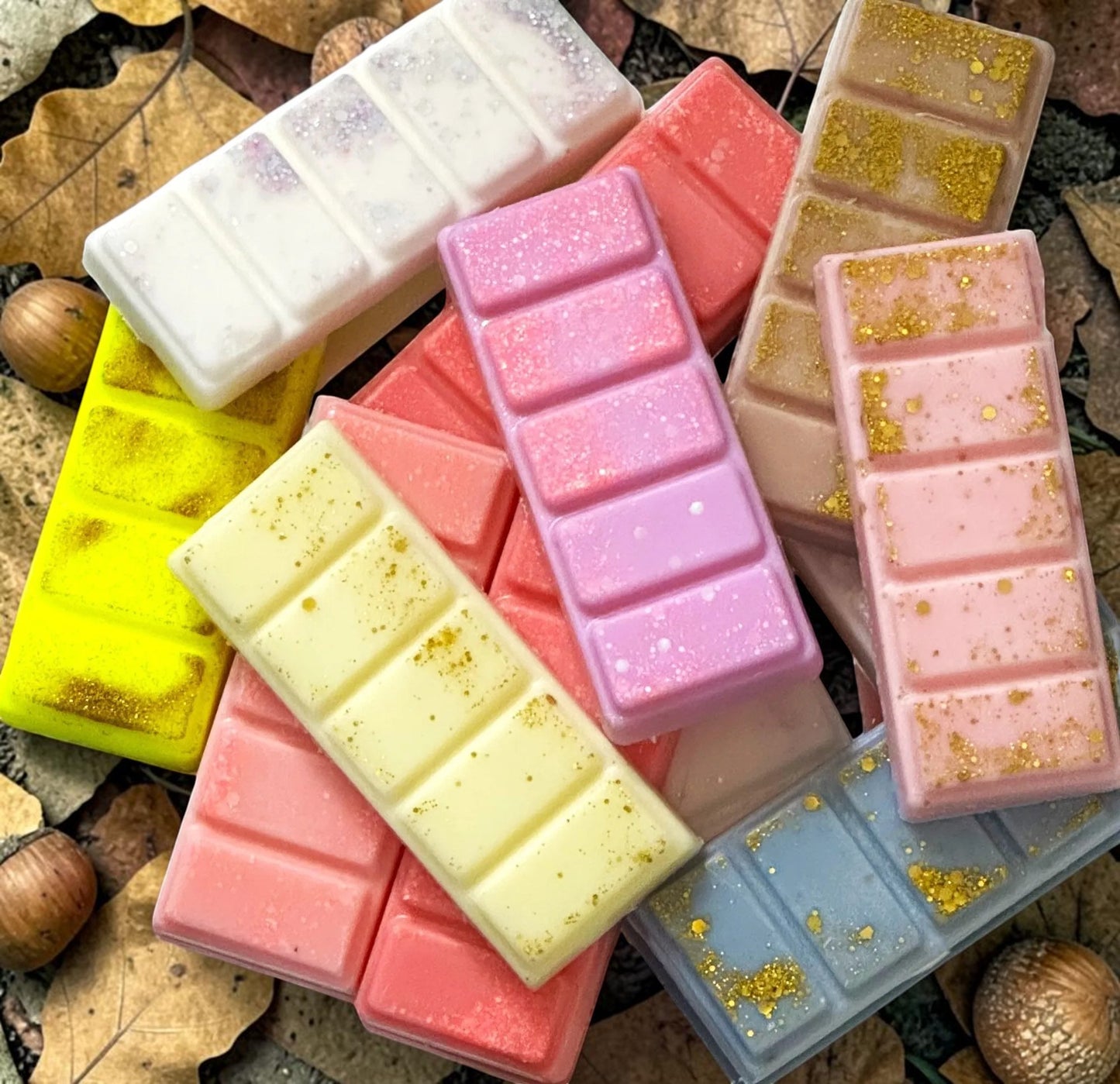 A collection of colorful wax melts shaped like chocolate bars, scattered on a bed of autumn leaves and acorns. The wax melts come in pastel shades of pink, purple, yellow, blue, and white, with some featuring sparkling glitter and others dusted with gold foil.