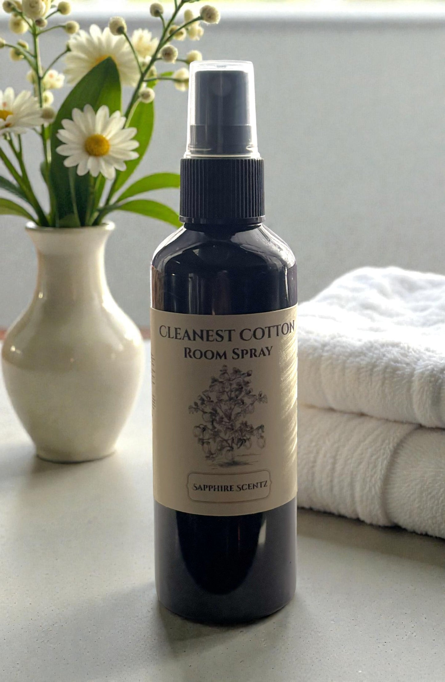 Room spray bottle labeled 'Cleanest Cotton' on a surface with flowers and towels in the background