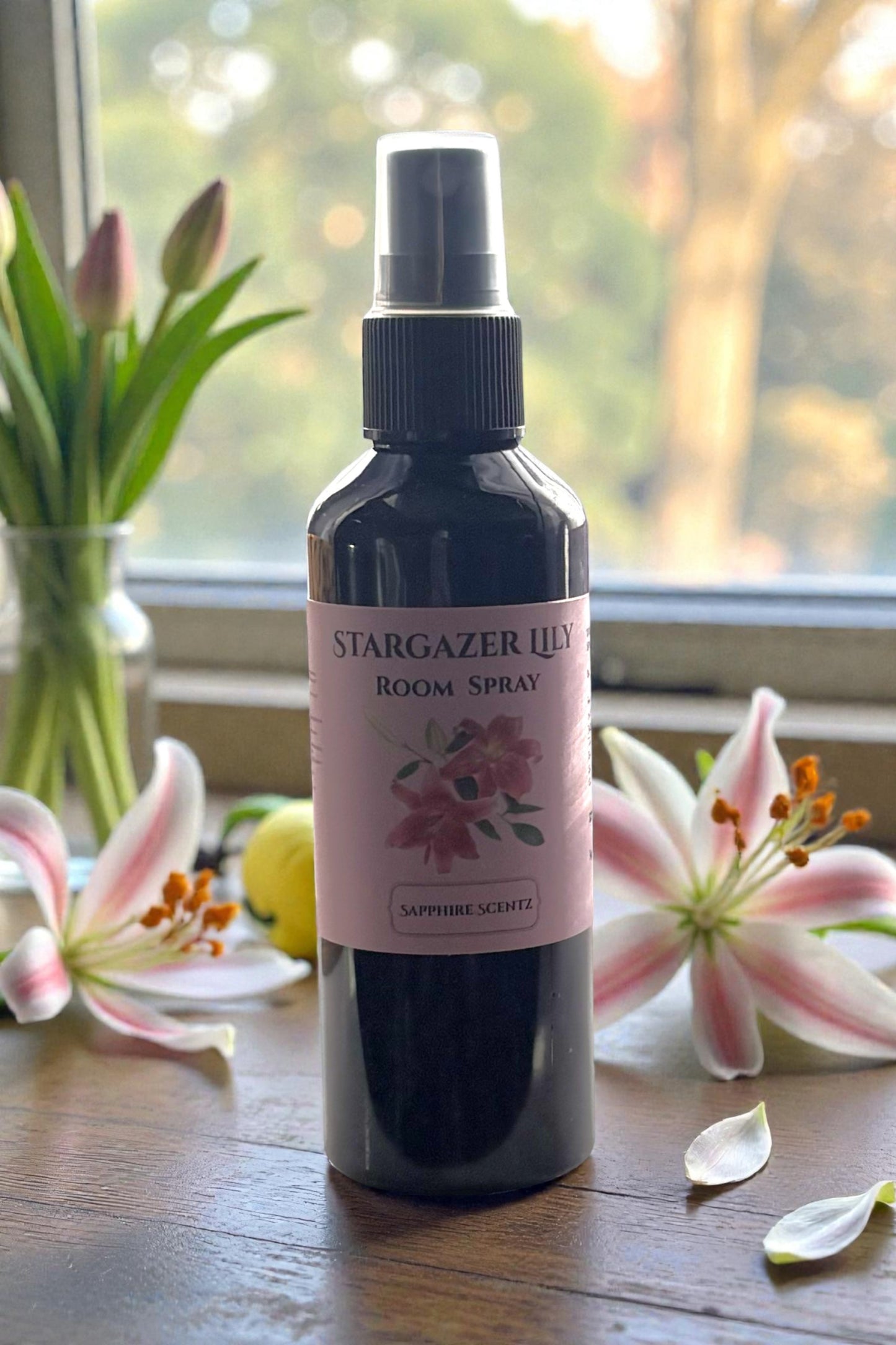 Room spray bottle labeled 'Stargazer Lily' on a wooden surface with flowers in the background.