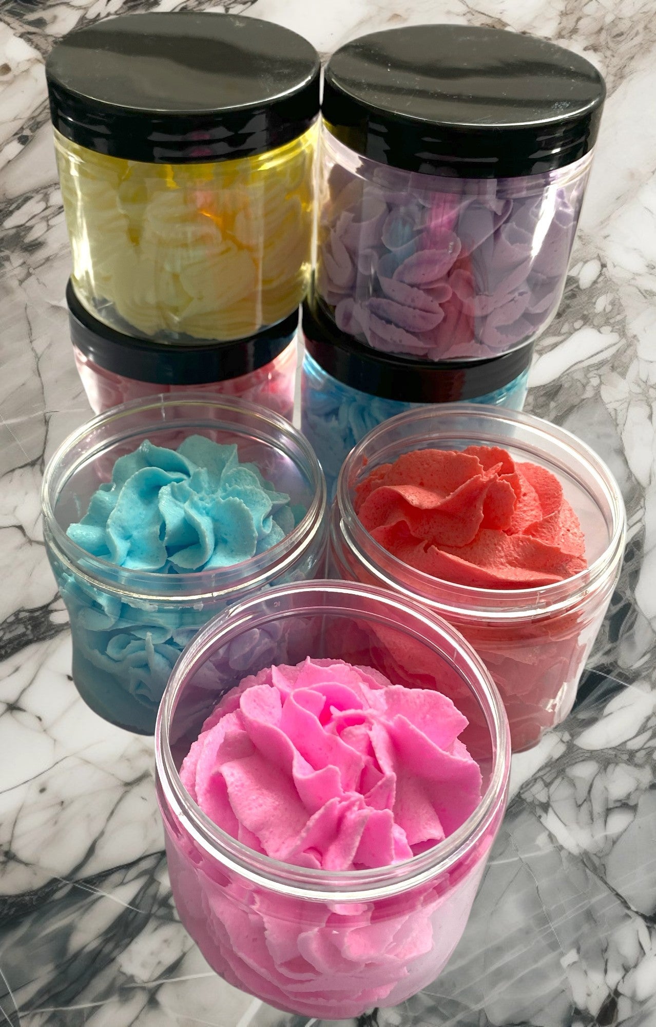 Multiple jars of whipped soap in various colors including yellow, pink, blue, and red, arranged on a marble surface.
