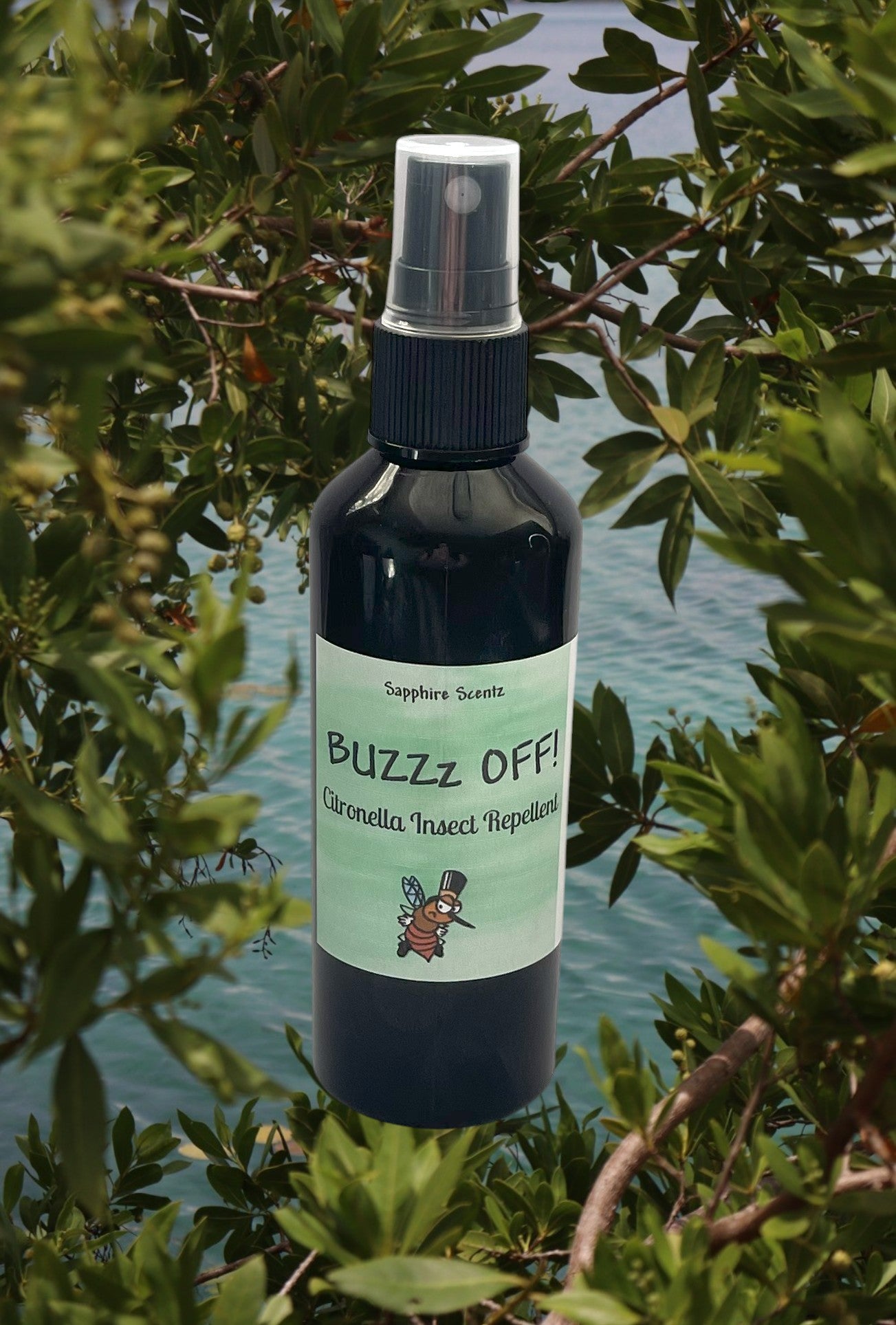 A black pump bottle of 'Buzz Off' citronella room and linen spray, labeled with white and green text, placed against a backdrop of greenery and water.