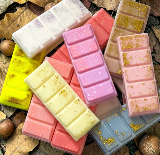 A collection of colorful wax melts shaped like chocolate bars, scattered on a bed of autumn leaves and acorns. The wax melts come in pastel shades of pink, purple, yellow, blue, and white, with some featuring sparkling glitter and others dusted with gold foil.