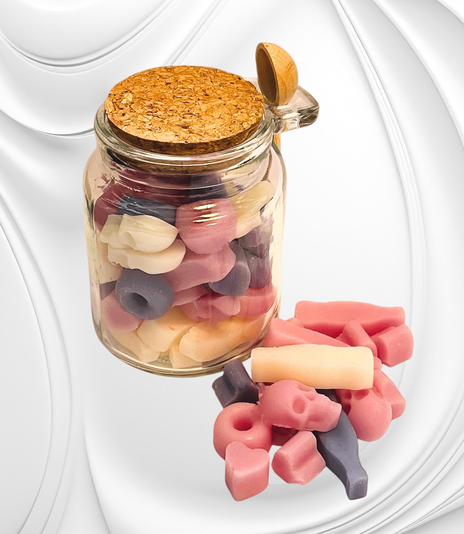 A clear glass jar with a cork lid, filled with multicolored wax scoopies, alongside a metal scoopie spoon. The scoopies are in shapes of hearts and rings, and the colors include pink, red, yellow, and blue.