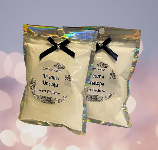 Two packets of 'Dreams Unstoppables Carpet Freshener' with a white label, black text, and decorative elements, tied with a black ribbon, against a sparkling background.