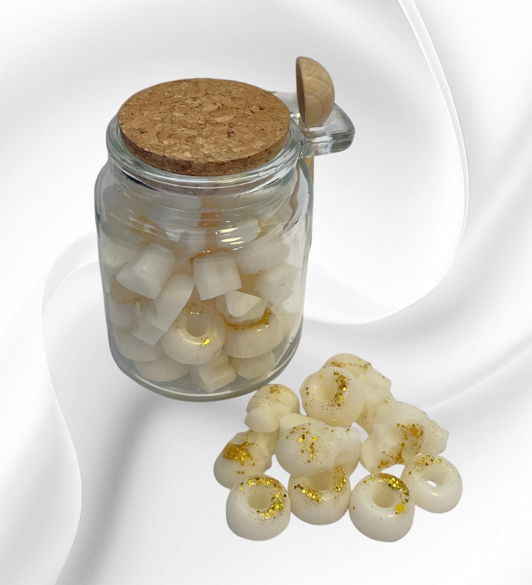 A clear glass jar with a cork lid, filled with white fluffy towel-shaped wax scoopies, with a metal scoop spoon beside it.