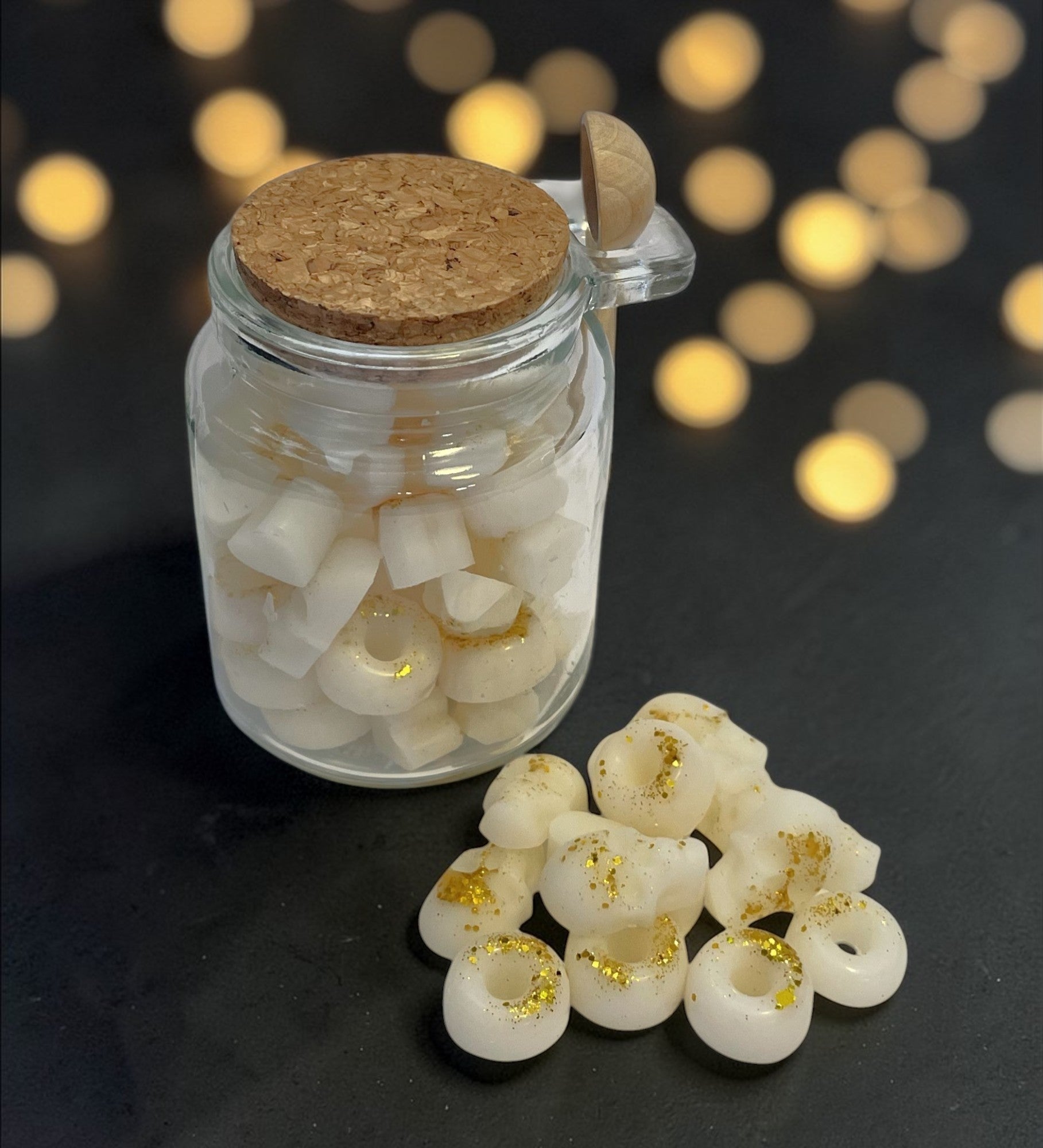 Jar of white donut-shaped candies with gold glitter on a dark surface with blurred lights in the background