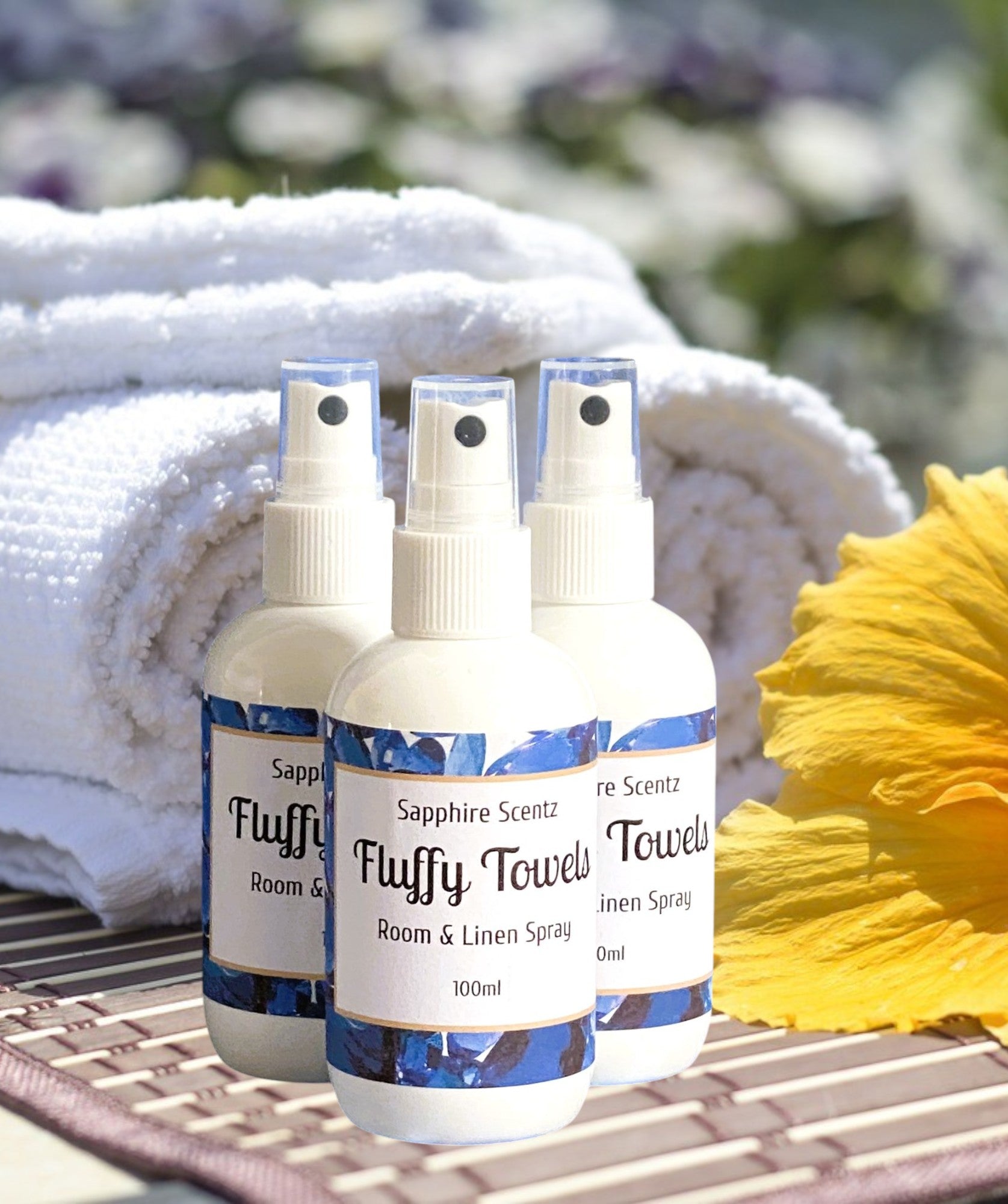 Three pump action room and linen sprays with labels 'Sapphire Scents Fluffy Towels', placed on a bamboo mat with a yellow flower and a white towel in the background.