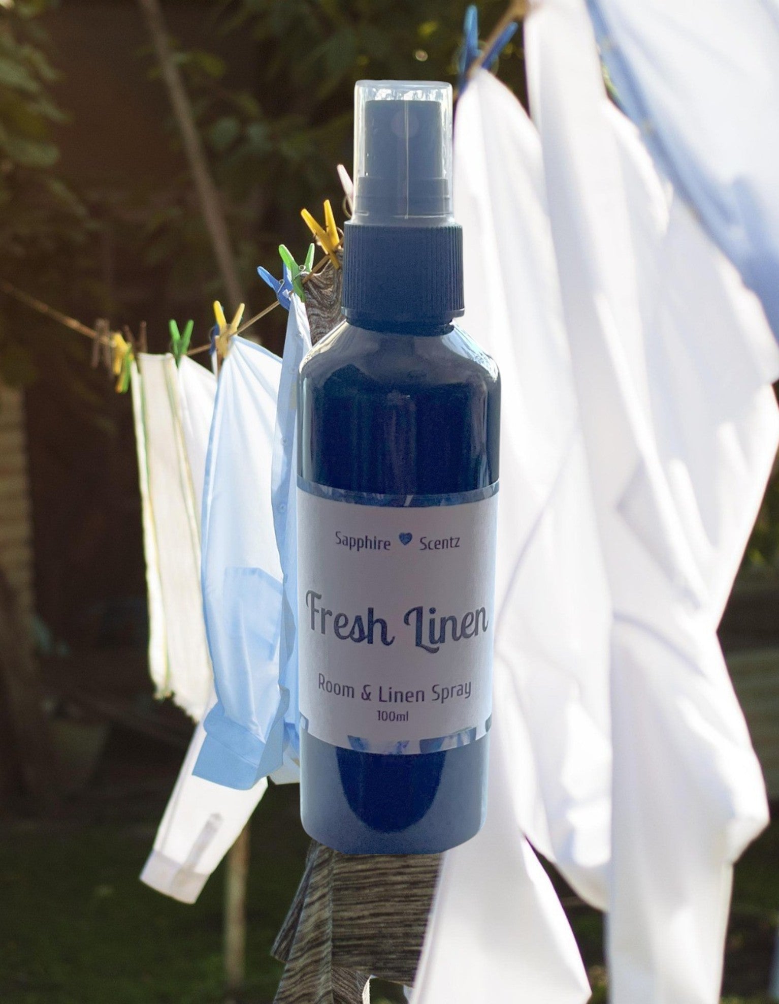 A blue bottle of 'Fresh Linen' room and linen spray with a pump dispenser, labeled and placed in an outdoor setting with clothes hanging in the background.