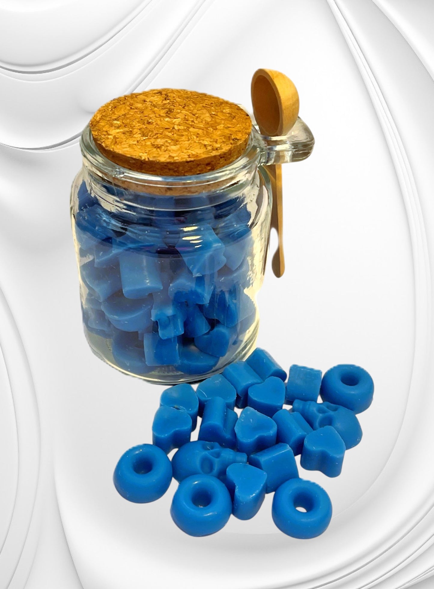 A clear glass jar with a cork lid, filled with blue wax scoopies, alongside a wooden scoopie spoon, with additional blue wax scoopies displayed in front.