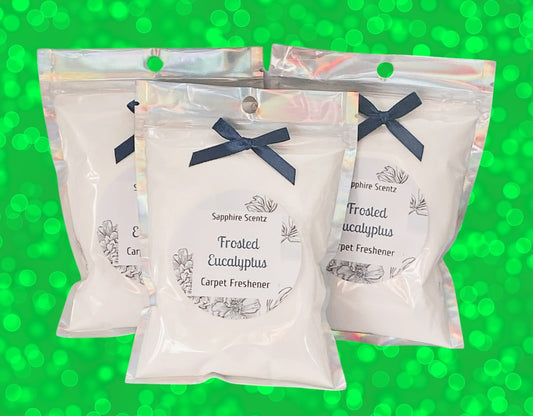 Three triangular packets of 'Frosted Eucalyptus' carpet freshener with white labels and blue ribbons, against a green background.