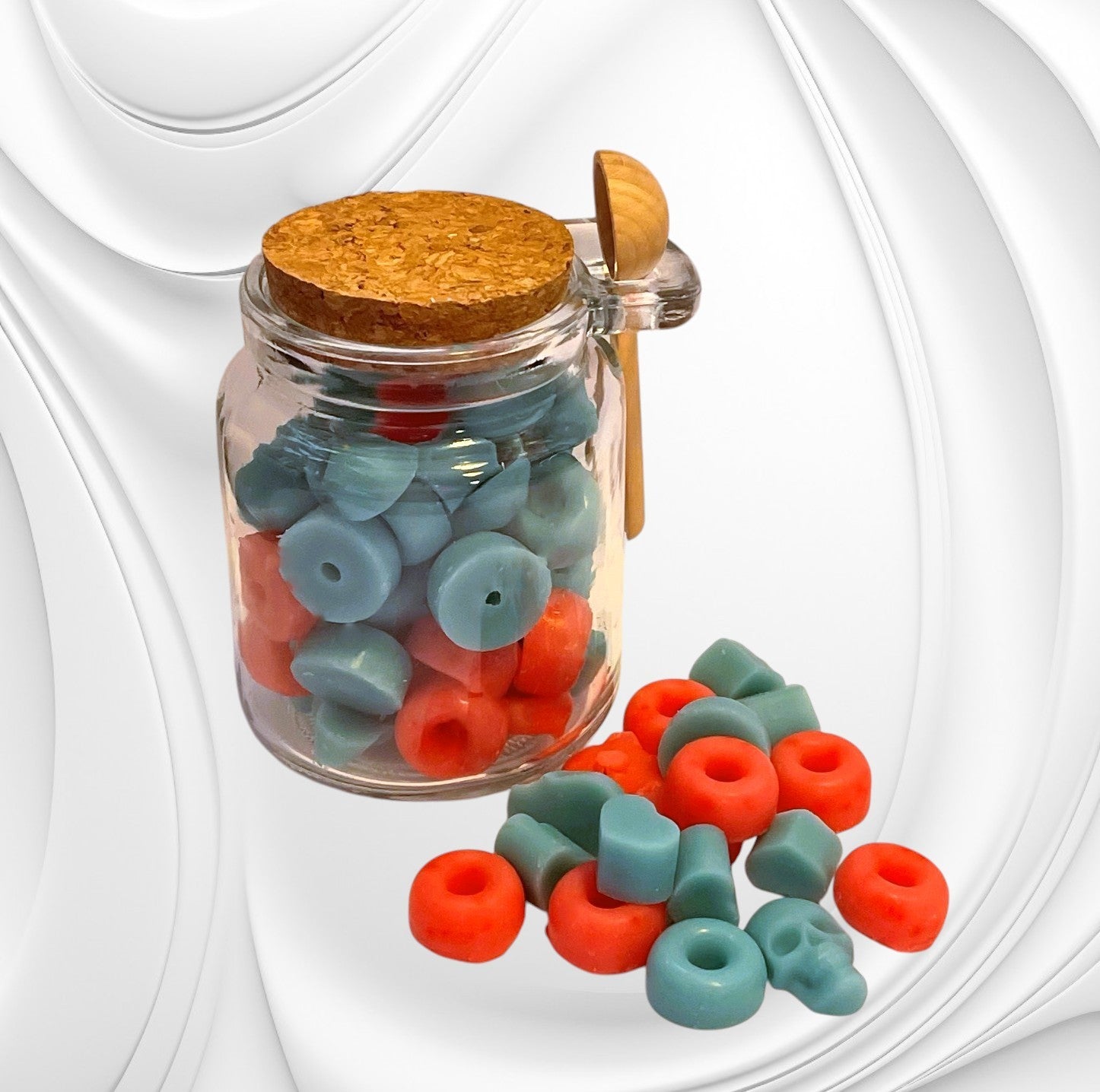 A clear glass jar with a cork lid, filled with blue and red wax scoopies, with a metal scoopie spoon beside it.