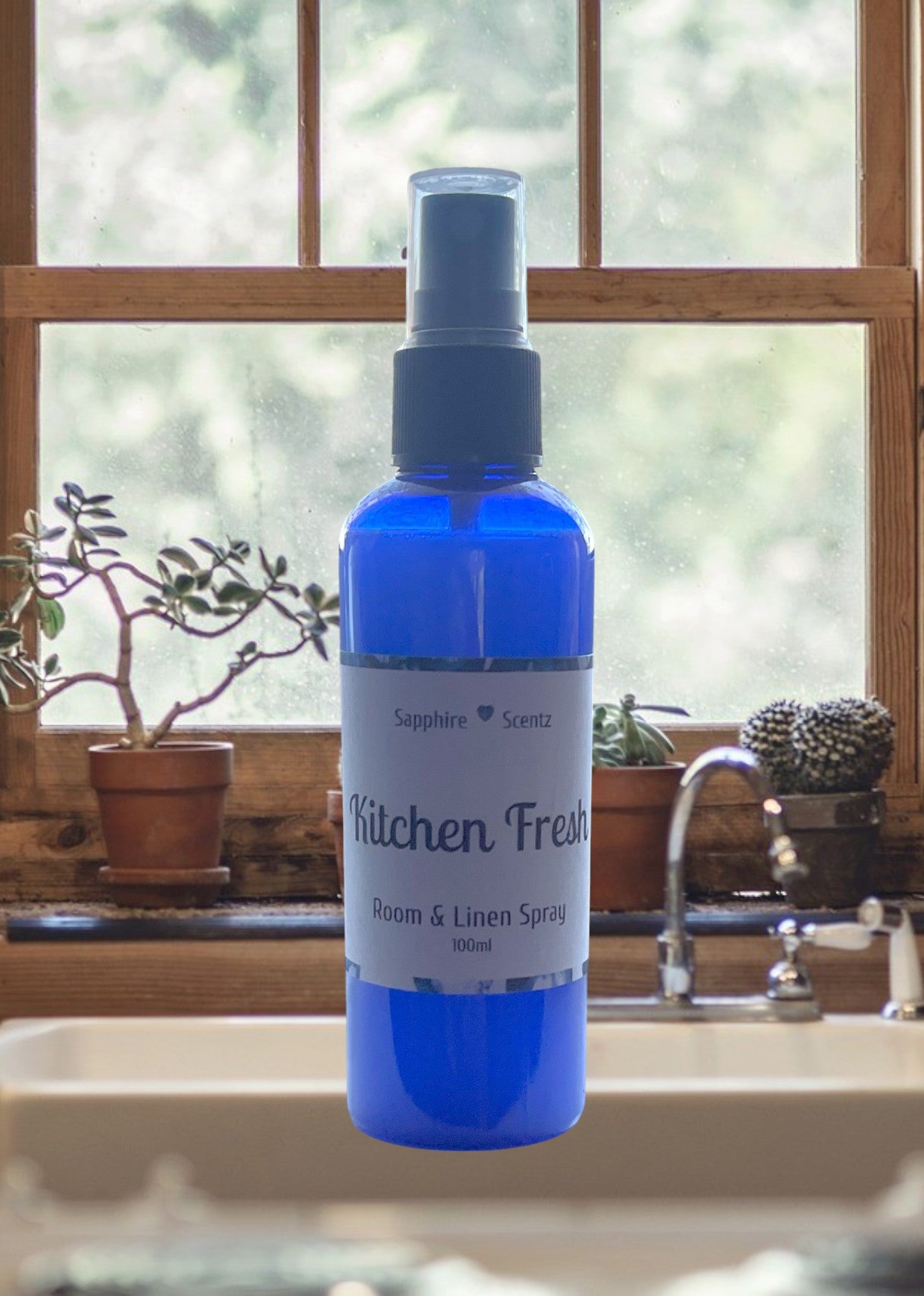 A blue room spray bottle labeled 'Kitchen Fresh' with a pump dispenser, placed on a windowsill with plants and kitchenware in the background.