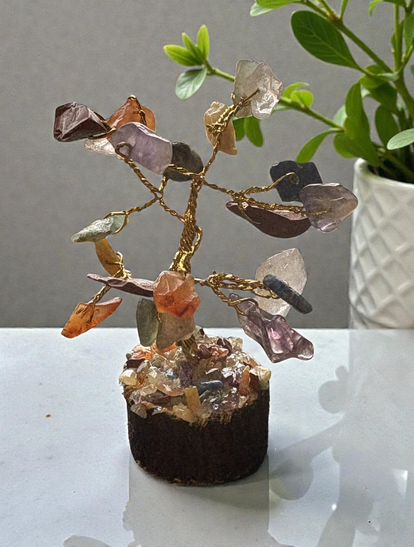 Crystal tree with multicolored stones on a white surface, with a plant in the background.