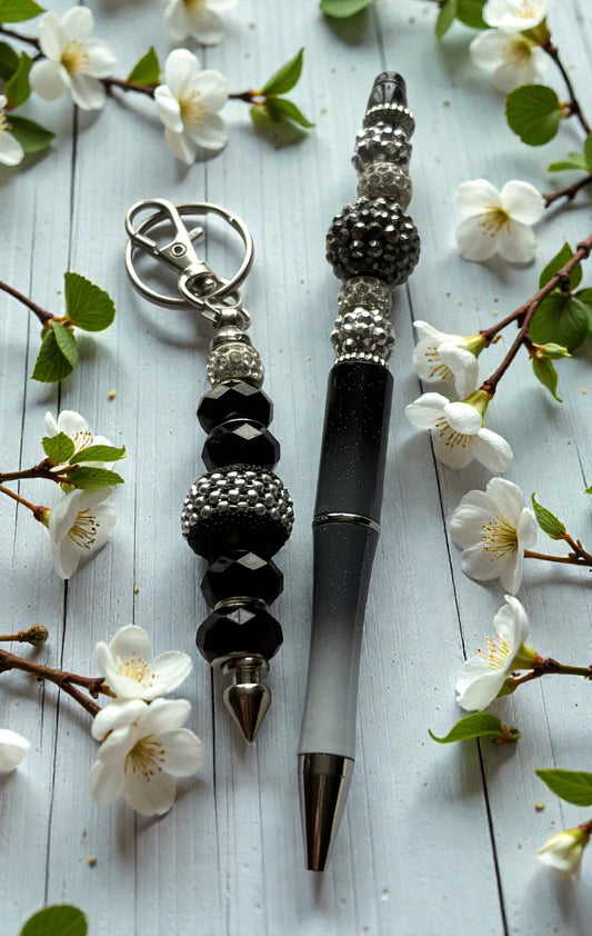 Decorative pen and keychain with black and silver beads on a light wooden surface with white flowers.