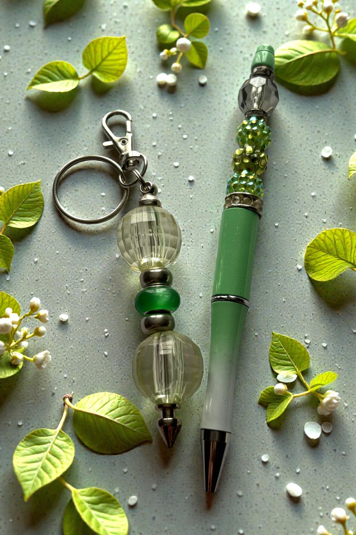 Green pen and keychain with decorative beads on a light gray background with leaves and flowers.