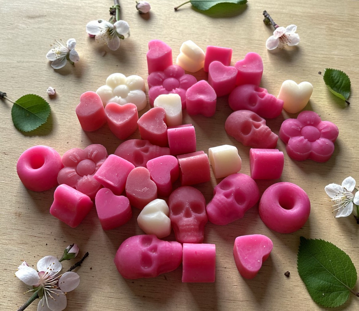 Pink and white wax melts shaped like hearts, flowers, and skulls on a wooden surface with leaves and flowers.