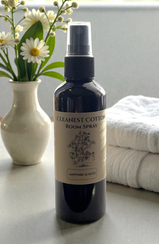 Room spray bottle labeled 'Cleanest Cotton' on a surface with flowers and towels in the background