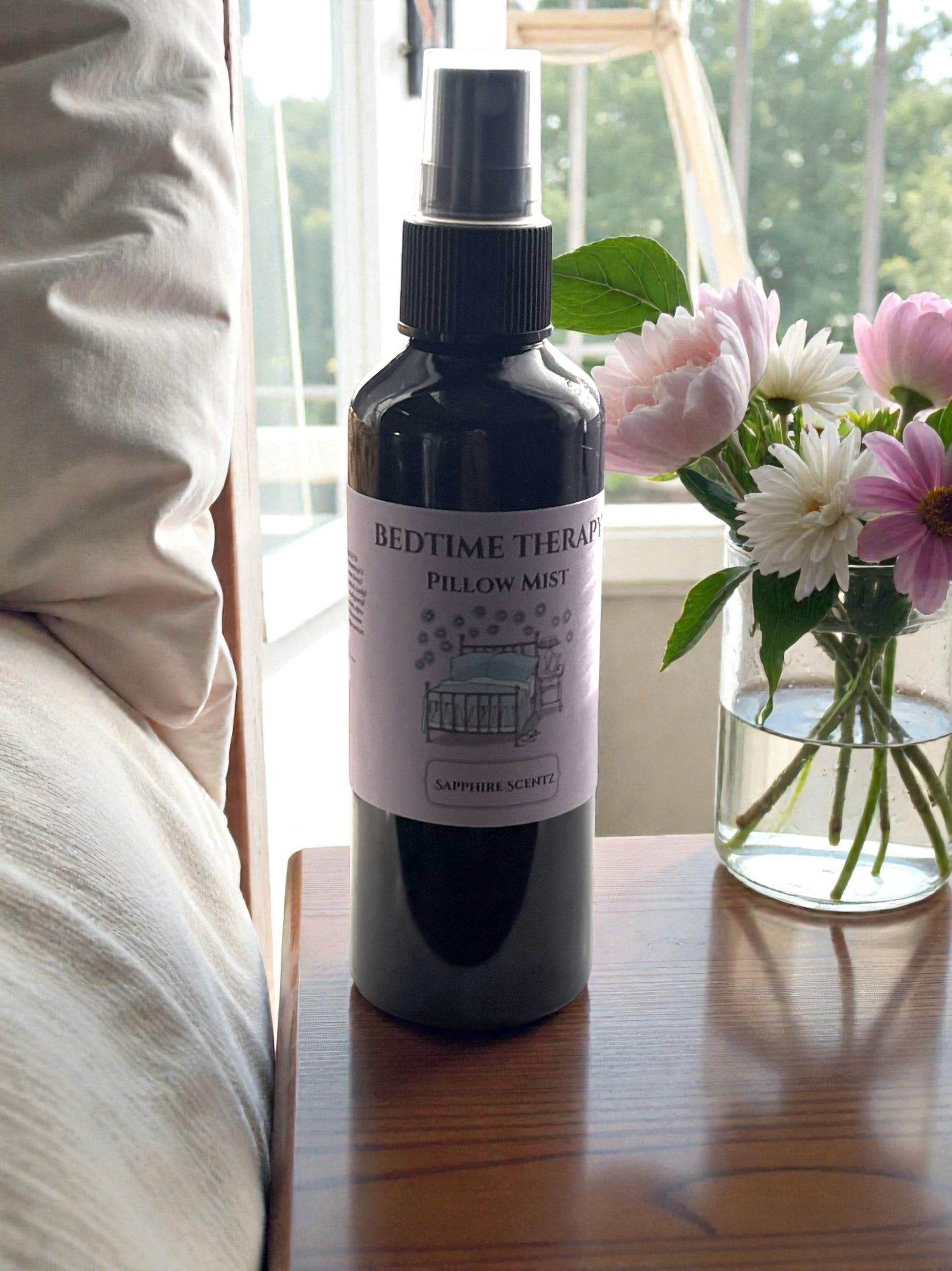 Bottle of 'Bedtime Therapy' pillow mist on a wooden surface with flowers in the background.
