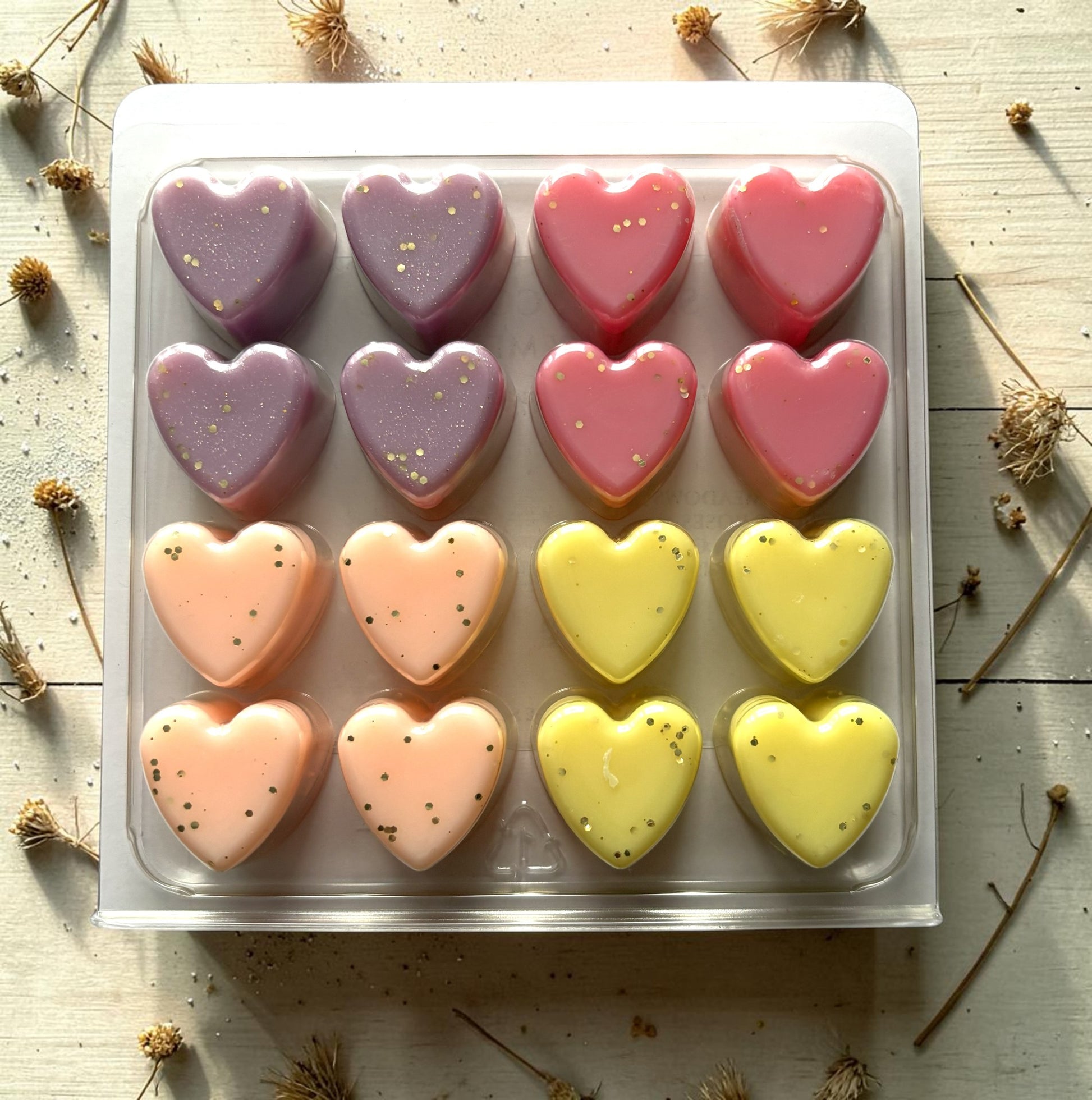Heart-shaped pastries in a clear plastic tray on a wooden surface with dried flowers.