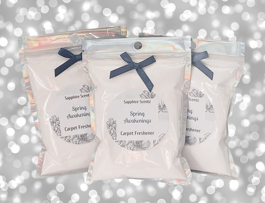 Three packets of 'Spring Awakenings - Carpet Freshener' with a white label and blue ribbon on each, placed against a glittery background.